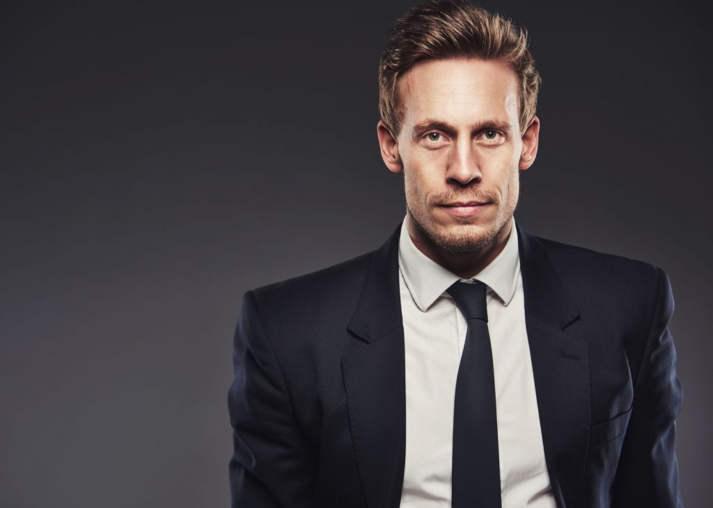 man in a suit and tie with dark grey background looking at  directly